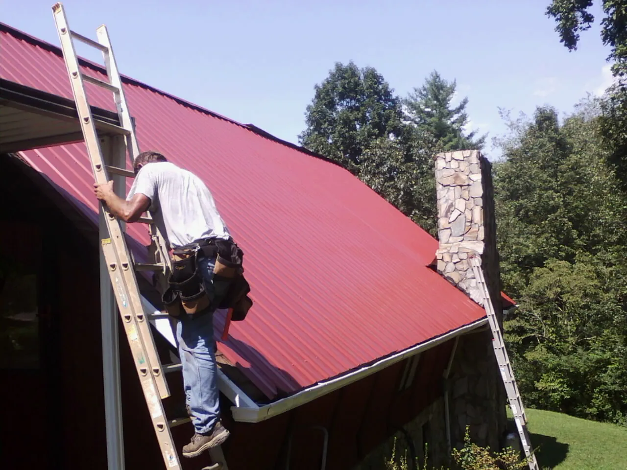 Roofer on a ladder beside a red metal roof and stone chimney — Soffit & Fascia Repair by Roofing Brothers in Ohio City